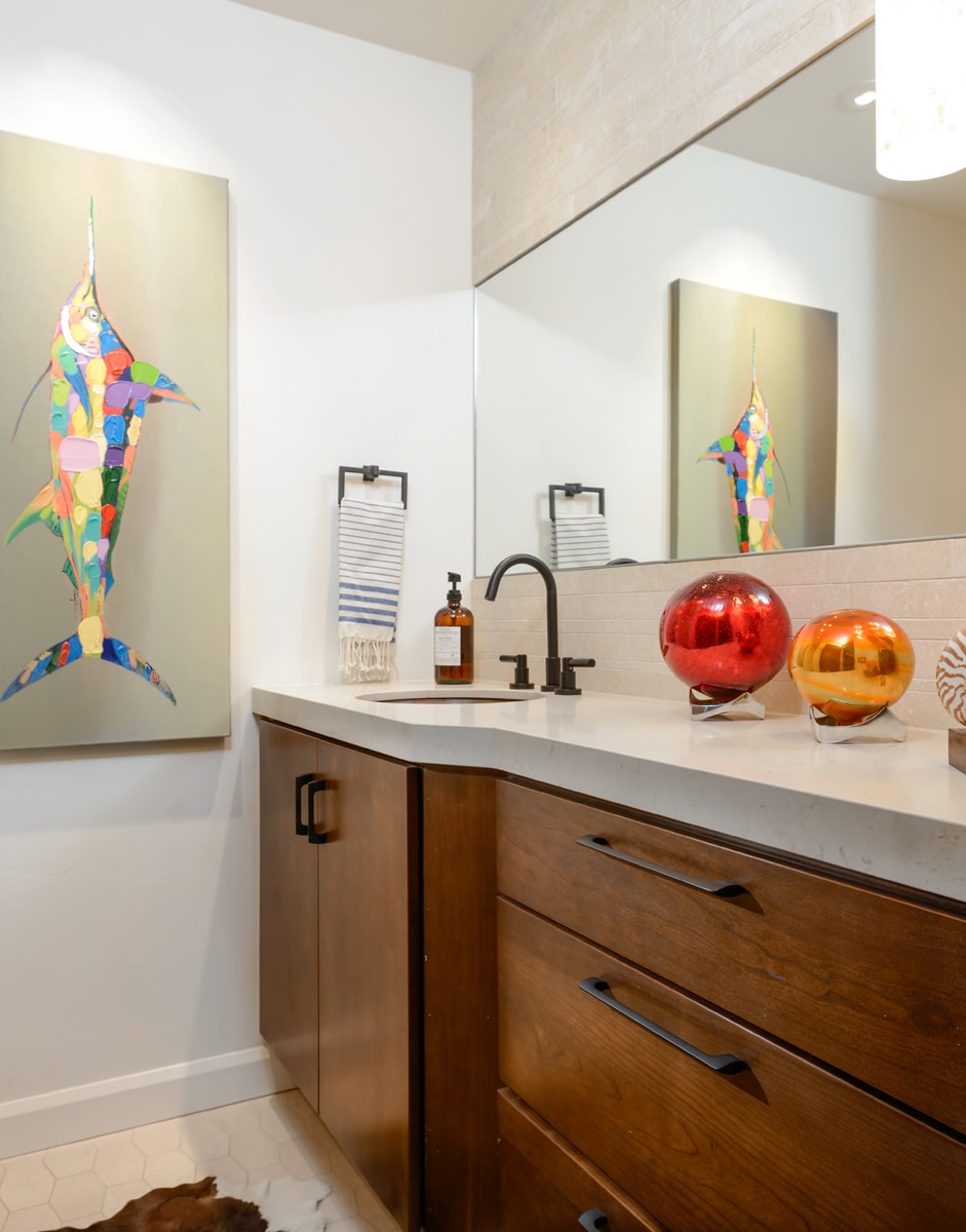 A warm wood bathroom vanity with red and orange accessories and a color fish painting on a white bathroom wall.